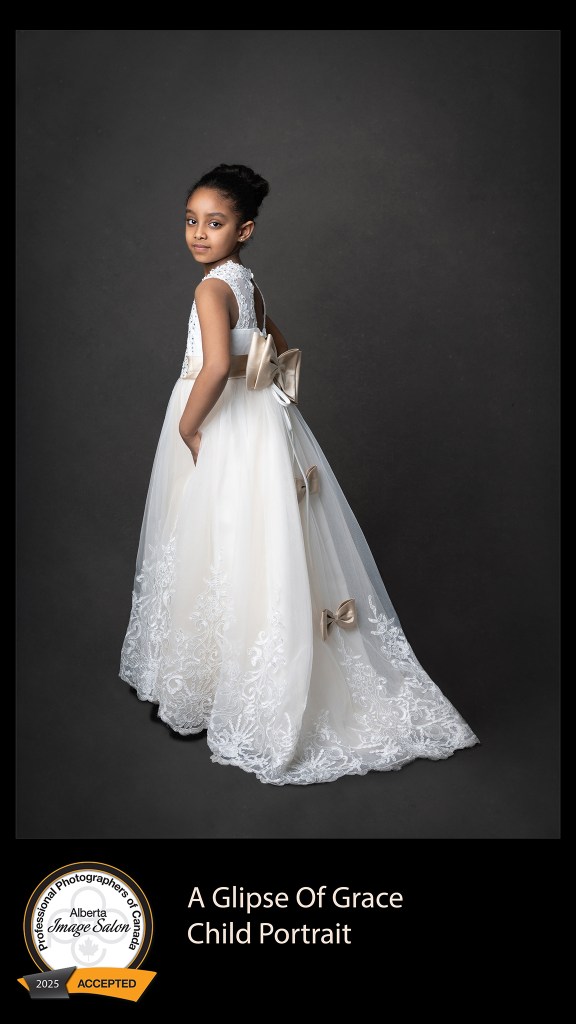 A studio portrait of a child looking back with a grey backdrop in a cream colored dress.